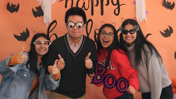 Un grupo de cuatro personas en disfraces de Halloween frente a un fondo naranja en el campus de la Burnaby North School.