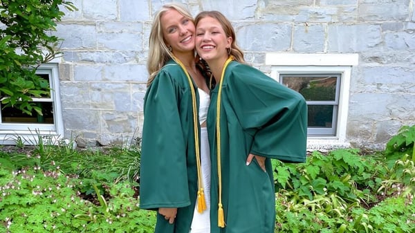 Dos estudiantes de la Burr and Burton Academy en togas de graduación verdes se abrazan frente a un edificio de piedra con un entorno verde.