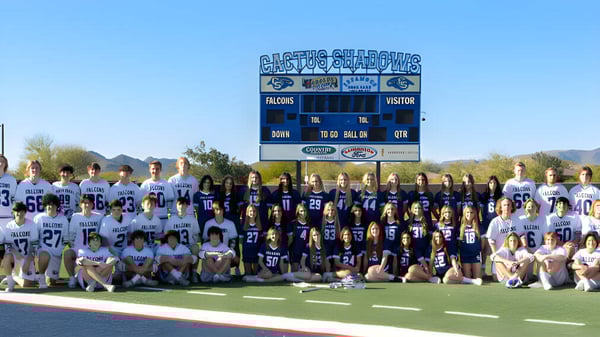 Jugadoras y jugadores de fútbol de la Cactus Shadows High School están en el campo frente a un marcador con montañas al fondo.