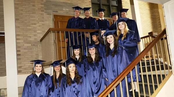 Un grupo de egresados de la Cair Paravel Latin School está de pie en togas azules en una escalera frente a un edificio de ladrillo.