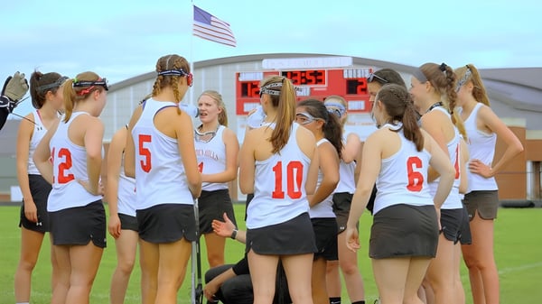 Un grupo de jóvenes atletas está en el campo de deportes de la Camden Hills Regional High School con una bandera americana al fondo.