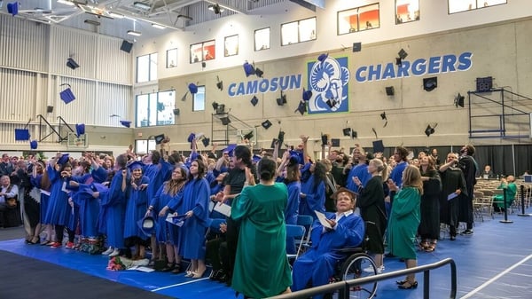 Un gran grupo de graduados en togas azules y verdes se reúne en un gimnasio en el campus de Camosun College.