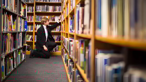 Una persona con un abrigo negro está en el estrecho pasillo entre altos estantes de libros en la biblioteca del Campbell College.