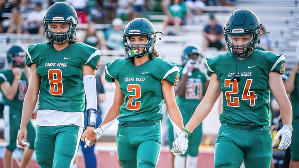 Tres jugadores de fútbol americano de la Campo Verde High School en camisetas verdes están en el campo frente a los espectadores.