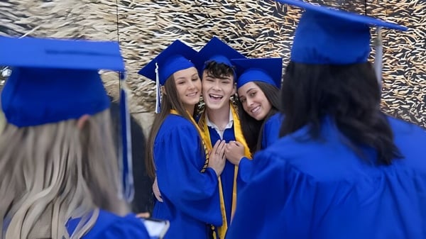 Los graduados de Canterbury High School celebran su graduación en togas y birretes azules frente a un fondo de árboles.