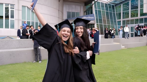 Dos graduadas de la Capilano University se abrazan frente a un edificio universitario con más graduados al fondo.