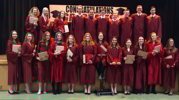 Un grupo de graduados de la Carbonear Collegiate High School está de pie en togas rojas en un escenario con un banner que dice 'Congratulations'.
