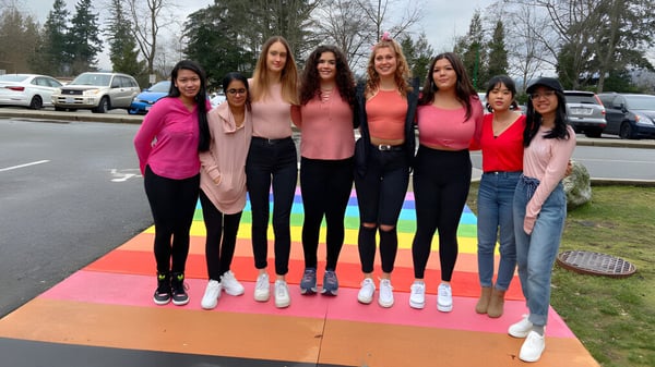 Un grupo de alumnas de la Cariboo Hill School está en una colorida terraza exterior frente a árboles y vehículos estacionados.