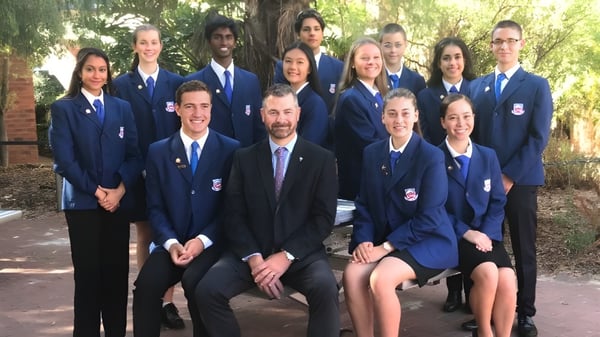 Un grupo de alumnos de la Carine Senior High School está al aire libre frente a un edificio de ladrillo con árboles de fondo.