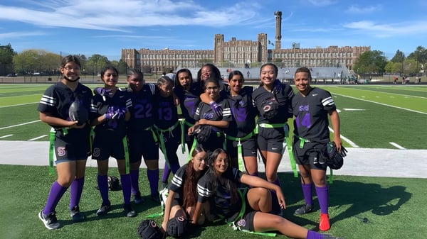 Un grupo de estudiantes en uniformes deportivos posan en el campo deportivo frente al edificio histórico de la Carl Schurz High School.