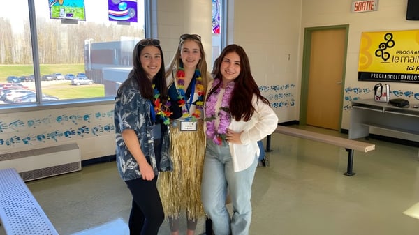 Tres estudiantes sonríen en el pasillo de la Carrefour de l’Acadie frente a casilleros y carteles escolares.
