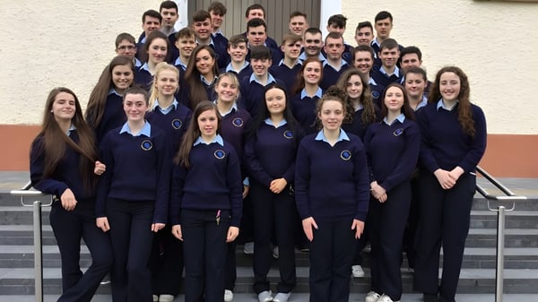 Un grupo de estudiantes en uniformes azul oscuro uniformes está frente a una pared blanca en el terreno de la Carrigallen Vocational School.