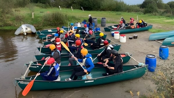 Estudiantes de la Carrigallen Vocational School están sentados en kayaks con chalecos salvavidas de colores en un río con densa vegetación.