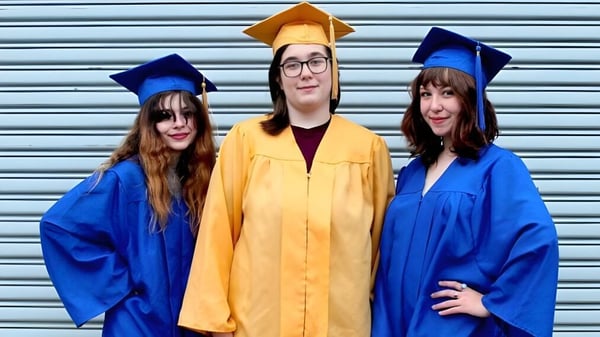 Tres personas con diferentes togas de graduación están frente a una persiana metálica en el campus del Castlepollard Community College.
