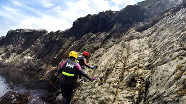 Un estudiante de la Castlerea Community School escala con un casco amarillo en un acantilado rocoso con vista al agua y las montañas.