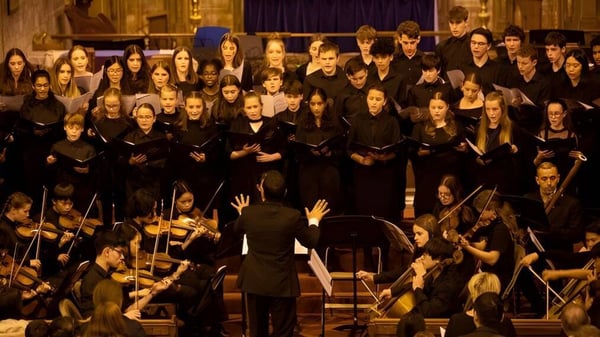 Un gran coro en túnicas negras canta en el escenario frente a una orquesta en la Caterham School.