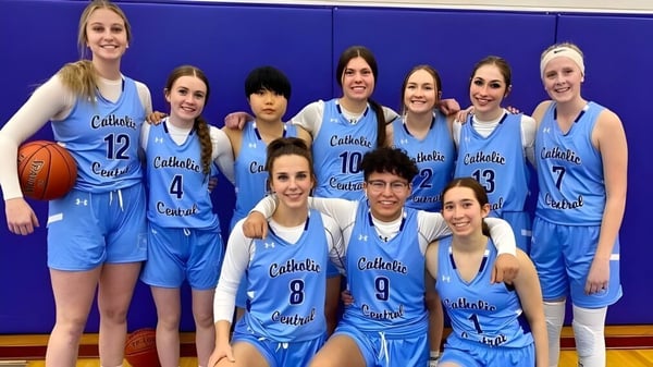 Un grupo de estudiantes en uniformes de baloncesto azules posan frente a una pared azul en el campus de la Catholic Central High School.