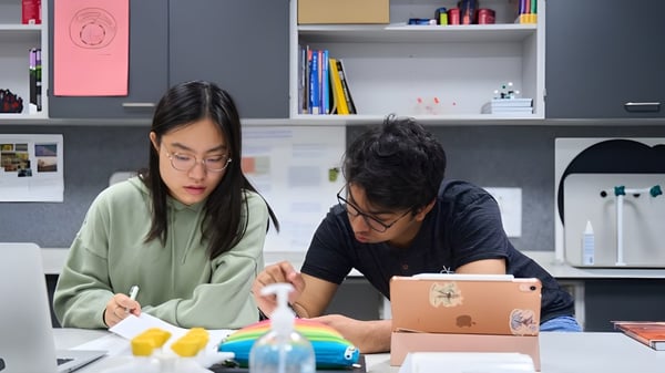 Dos personas trabajan juntas en un proyecto en una sala con estantes llenos de libros y materiales de oficina en el campus de CATS Cambridge.
