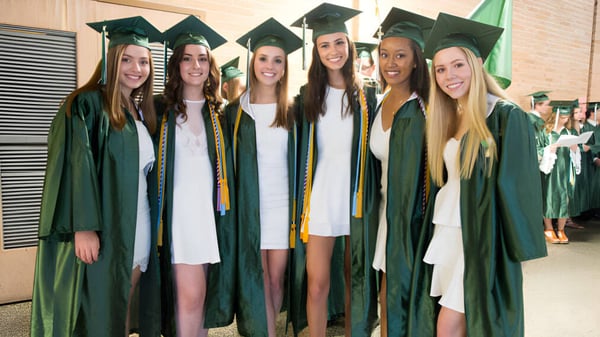 Un grupo de jóvenes mujeres en togas de graduación verdes celebra su graduación frente a una pared en el campus de la Cavendish Road State High School.