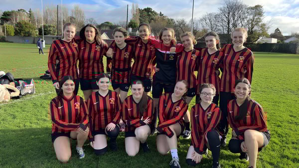 Un grupo de jóvenes futbolistas de CBS Westland Row posan en un campo de césped con árboles y edificios de fondo.