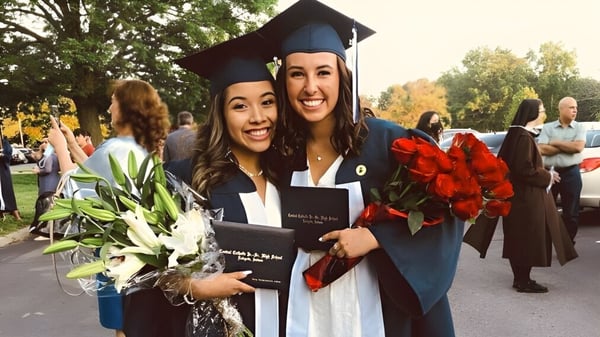 Dos estudiantes de la Central Catholic Jr.-Sr. High School en ropa de graduación sostienen flores y sonríen frente al fondo de otros graduados.