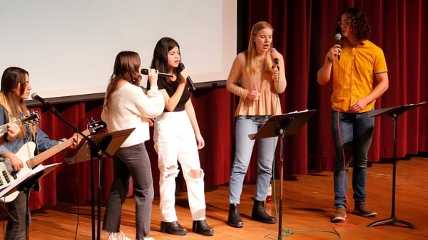 Un grupo de estudiantes de la Central Christian School ensaya en el escenario frente a una cortina roja con micrófonos.