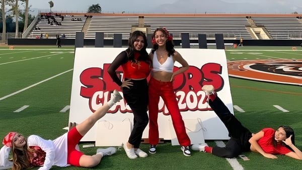 Dos estudiantes de la Chaffey High School posan frente a un cartel del Sports Club 2022 en el campo deportivo con un estadio de fondo.