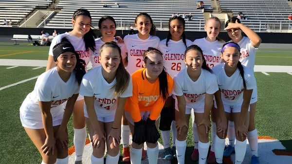 Un grupo de jóvenes jugadoras de fútbol posan juntas en el campo deportivo de la Chaffey High School con un estadio de fondo.