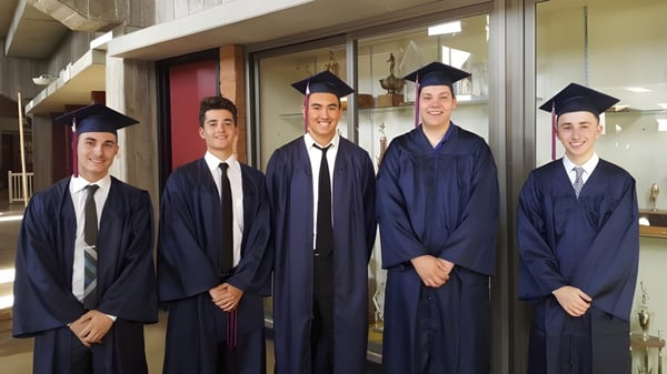 Un grupo de cinco estudiantes en gorros de graduación está de pie frente a una pared de vidrio en el campus del Chandler Unified School District.