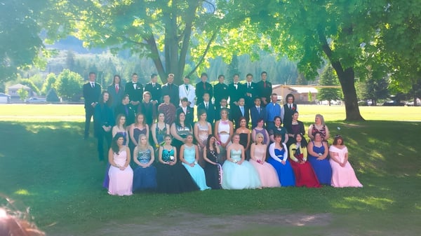 Un gran grupo de estudiantes de la Charles Bloom Secondary School se reúne al aire libre en un campo con árboles de fondo.