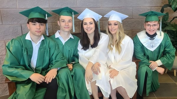 Un grupo de cinco graduados en togas verdes posan frente a una pared de ladrillo en The Charles Finney School.