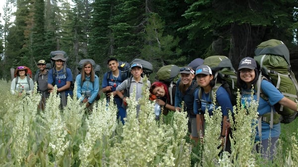Un grupo de estudiantes de la Charterhouse School está vestido para caminar en una pradera con hierba alta y flores silvestres frente a un denso bosque.
