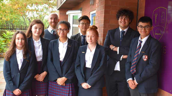 Un grupo de estudiantes de la Chase Grammar School está juntos frente a un edificio de ladrillo con plantas al fondo.