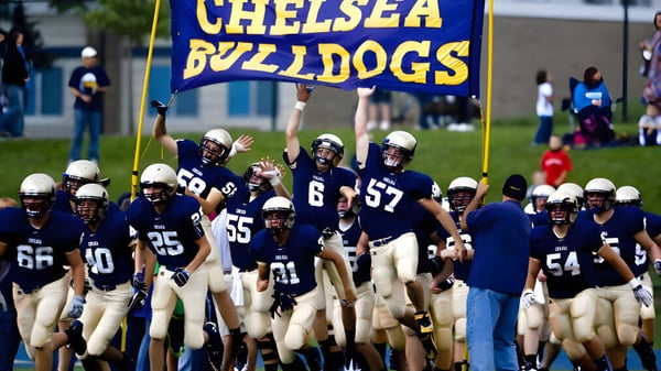 Estudiantes de Chelsea High School están en uniformes de fútbol en el campo con espectadores al fondo.