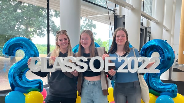Tres estudiantes sostienen un gran cartel con CLASS OF 2022 frente a un edificio en el campus de Cheshire College South & West.