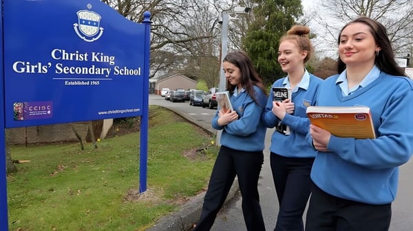 Tres estudiantes están frente a un cartel de la Christ King Secondary School con árboles y césped de fondo.