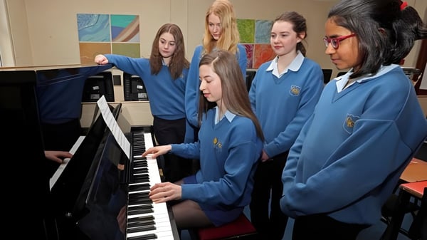 Estudiantes de la Christ King Secondary School en uniformes azules tocan el piano juntos frente a coloridas obras de arte.