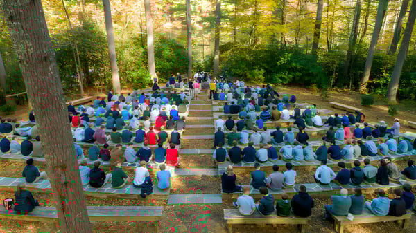 Un gran grupo de personas está sentado en bancos de madera en el bosque de la Christ School.