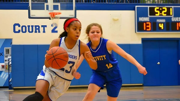 Dos estudiantes de la Christian Central Academy compiten en la cancha de baloncesto por el balón frente a un marcador.