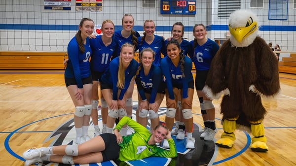 Un grupo de jugadoras de voleibol en camisetas azules posan juntas en la cancha de baloncesto de la Christian Life School con una mascota amarilla.
