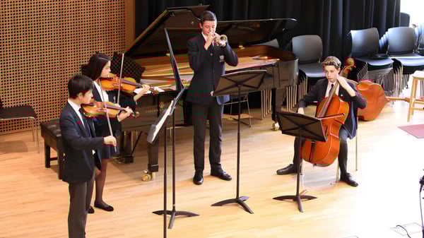 Estudiantes de la Claremont School actúan vestidos formalmente con diferentes instrumentos musicales en un escenario frente a una pared de paneles de madera.