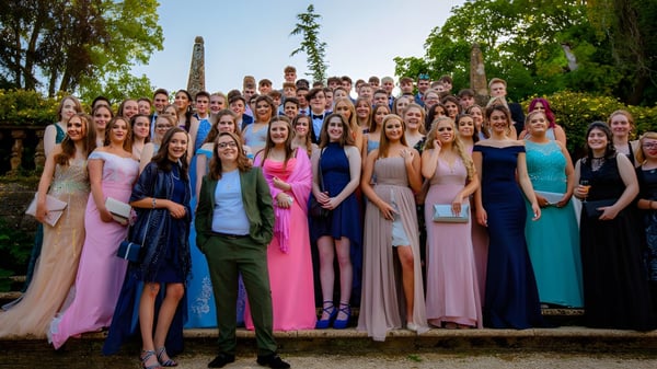 Un grupo de jóvenes mujeres en vestidos elegantes está juntas frente a un paisaje en el campus de la Claremont School.