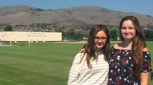 Dos estudiantes están en un campo cubierto de hierba con vista a las montañas en el terreno de la Clarence Fulton Secondary School.