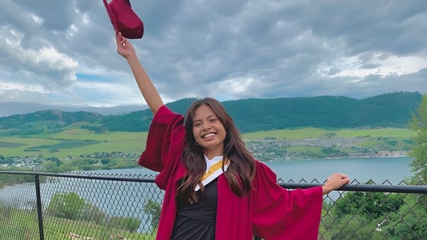Una joven en toga de graduación roja está en el balcón con vista a las montañas y un lago en Clarence Fulton Secondary School.