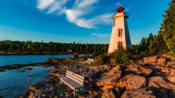 Un faro se encuentra en un acantilado rocoso frente a un lago tranquilo con un bosque al fondo en el terreno de la Clarke Road Secondary School.