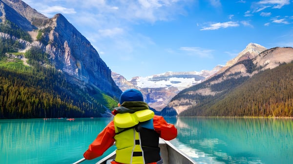 Una persona con colorido equipo de exterior está en un bote en el lago frente a montañas cubiertas de nieve cerca de la Clayton Heights Secondary School.
