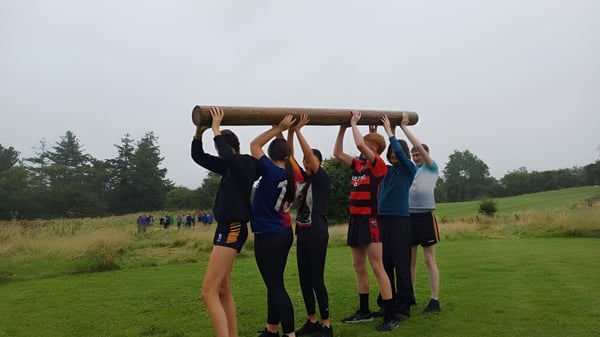 Estudiantes del Clonfert College levantan juntos un gran tronco en un prado con árboles.