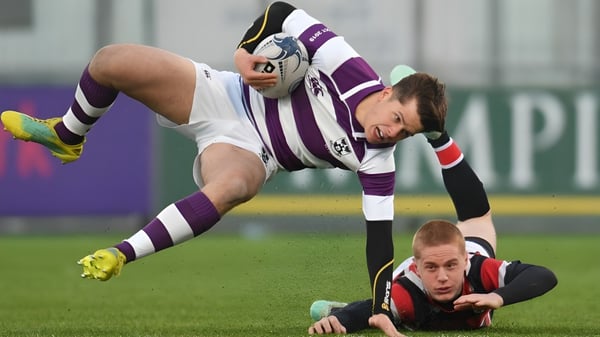 Un jugador con una camiseta a rayas morado y blanco salta sobre un jugador con una camiseta roja en el campo del Clongowes Wood College.