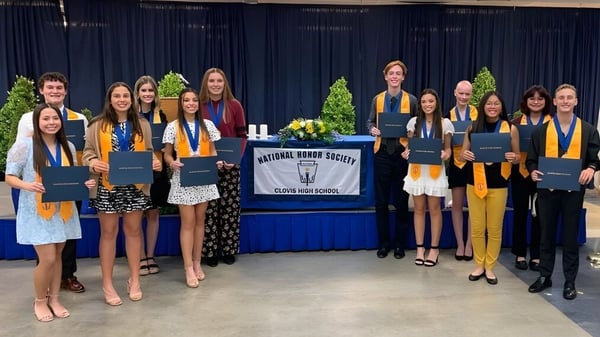 Un grupo de jóvenes mujeres en vestimenta de graduación se encuentra frente a un banner de la National Honor Society en el campus de la Clovis High School y sostiene sus diplomas.