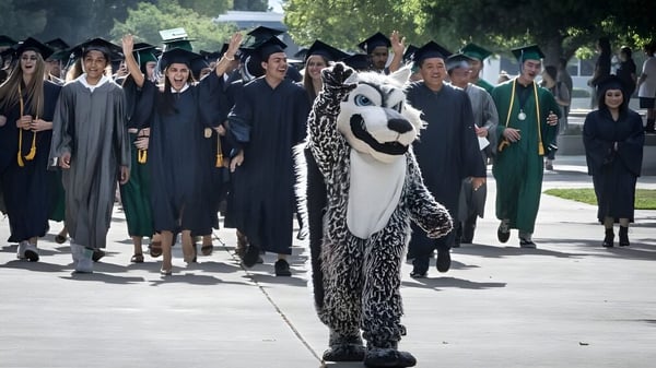 Un grupo de graduados del Clovis Unified School District lleva vestimenta académica y camina por un sendero del campus con árboles.